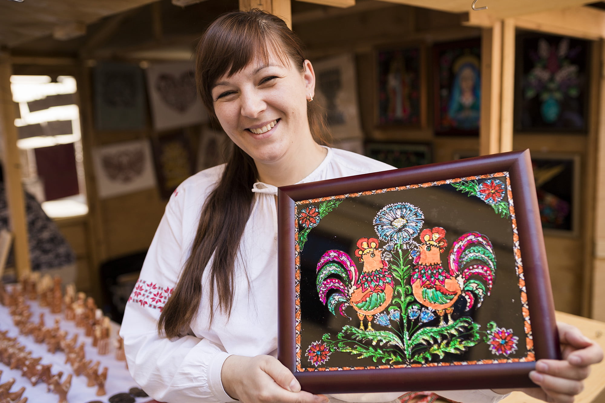 [Uśmiechnięta kobieta trzyma w ręku obraz w ramce przedstawiający kolorowe kury i kwiaty.]