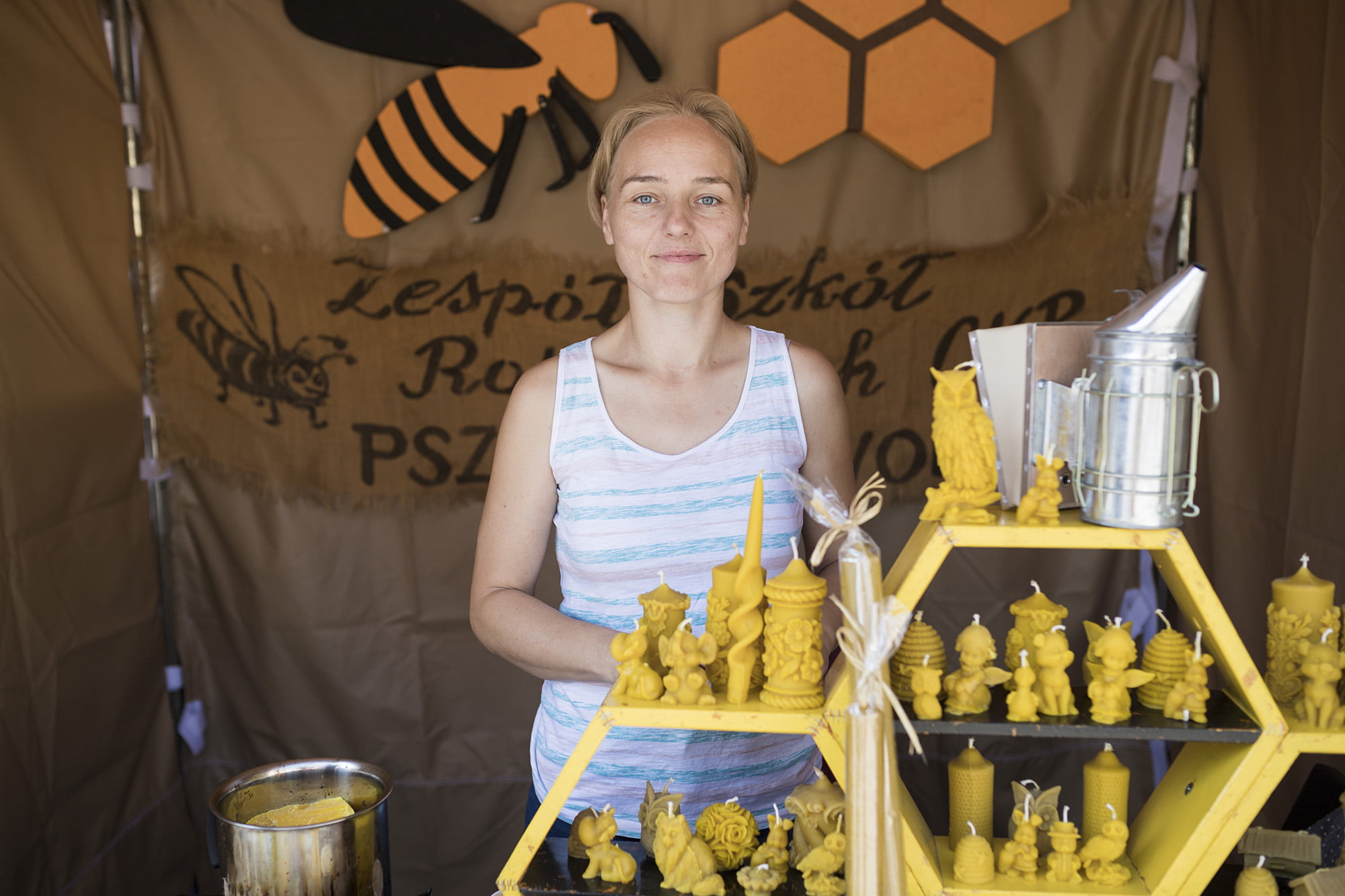 [Zbliżenie na stoisko pszczelarzy. Uśmiechnięta kobieta przed nię na półkach świece z wosku pszczelego.]