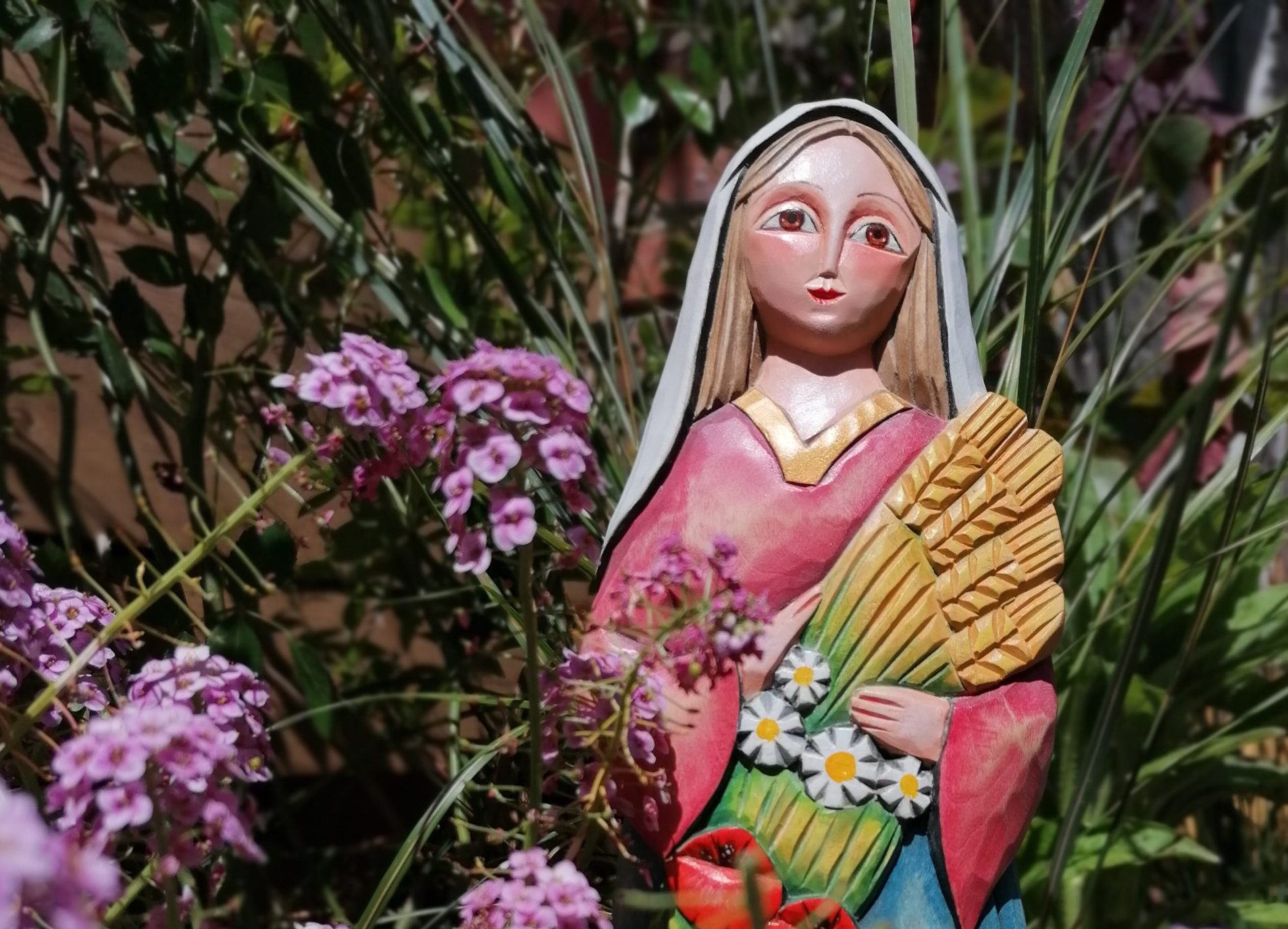 Drewniana rzeźba ukazująca Matkę Boską trzymającą kłosy zbóż i kwiaty. Rzeźba ustawiona wśród roślin i delikatnych, różowych kwiatostanach.