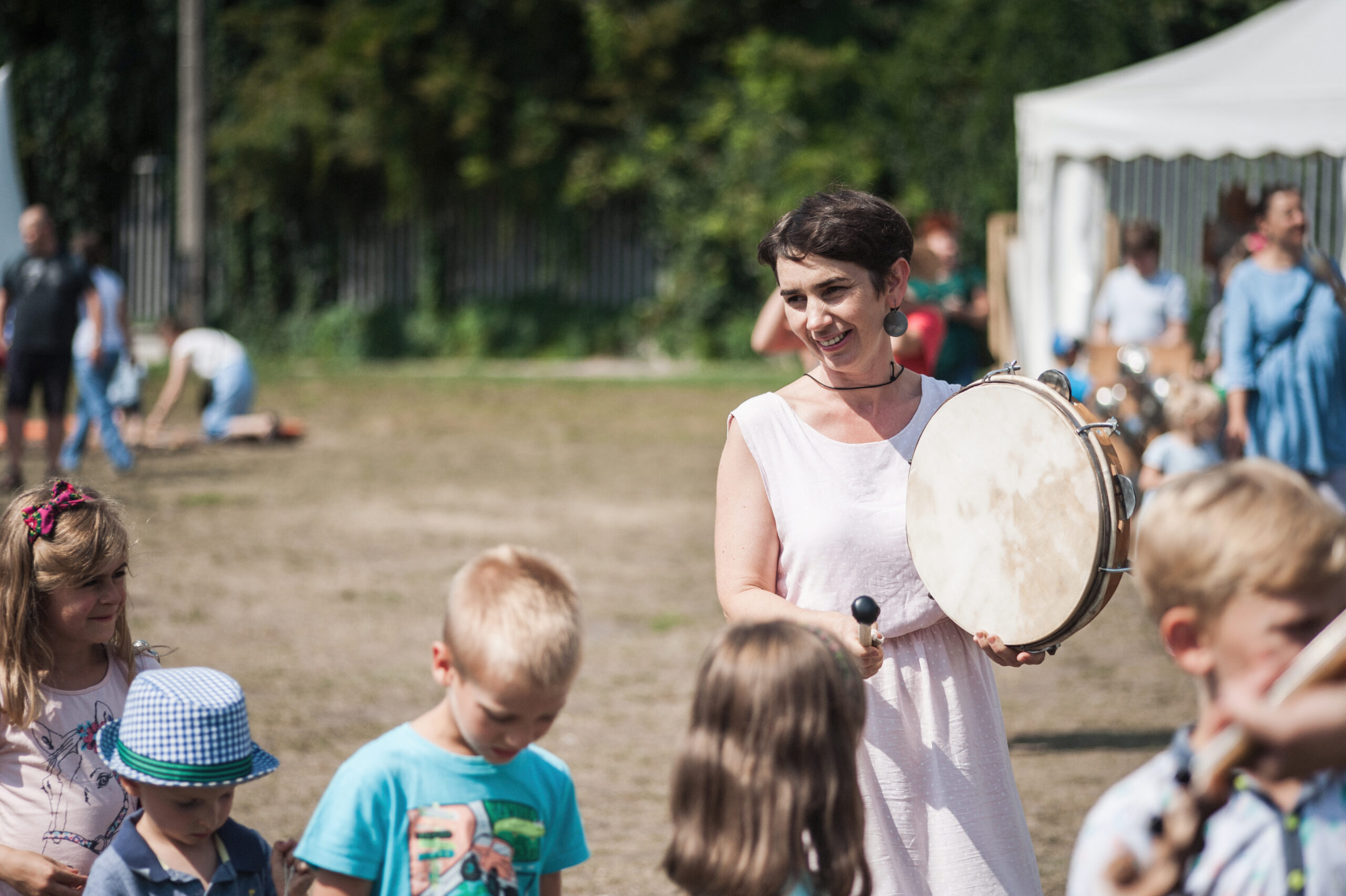 Marta Urban-Burdalska, uśmiechnięta kobieta z krótkimi czarnymi włosami trzyma w rękach bęben i pałeczkę, za pomocą której gra. Przed kobietą stoją dzieci.
