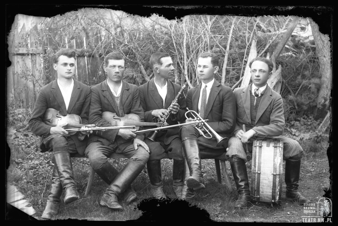 an old black and white posed photo showing five men sitting on a bench. Each is holding a instrument. We see two violins, a clarinet, a trupet and a drum - baraban