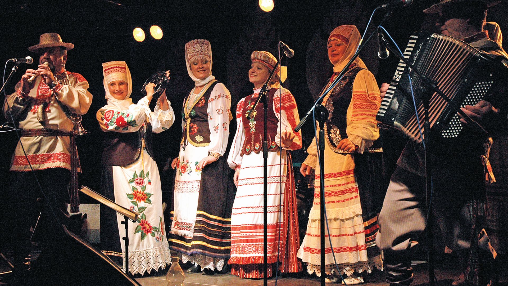 [Scena festiwalu. Widzimy cztery kobiety w tradycyjnych strojach białoruskich Po ich bokach stopją mężczyźni grający na instrumentach - akordeonie i flecie. Mężczyźni w strojach tradycyjnych]