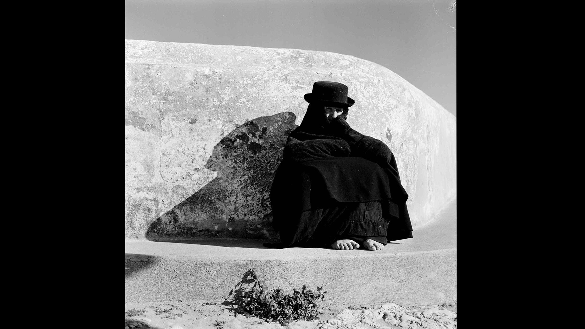 Fotografia czarno-biała. Kobieta w burce i kapeluszu oparta o skałę.